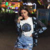 Young female model with long straight dark hair wearing high-waisted distressed denim shorts and a blue floral cardigan over a premium white graphic streetwear t-shirt featuring a detailed electric blue Aquarius lightning warrior design by Essentia Collective at an outdoor carnival at night