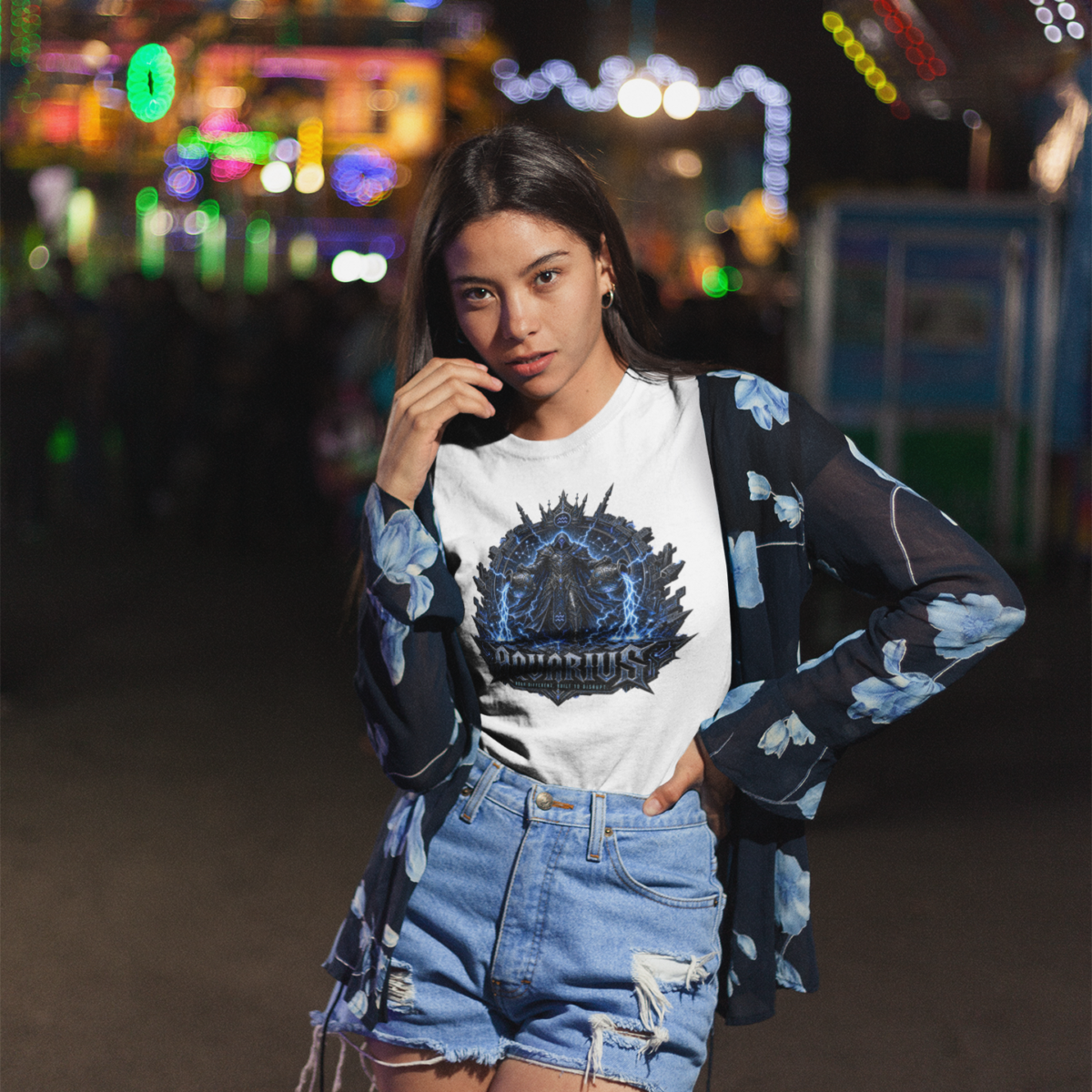 Young female model with long straight dark hair wearing high-waisted distressed denim shorts and a blue floral cardigan over a premium white graphic streetwear t-shirt featuring a detailed electric blue Aquarius lightning warrior design by Essentia Collective at an outdoor carnival at night