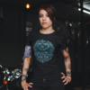 Heavily tattooed female model with an arm band bandana wearing a premium black graphic streetwear t-shirt featuring a detailed dark teal Pisces crest design by Essentia Collective inside a gritty industrial garage.