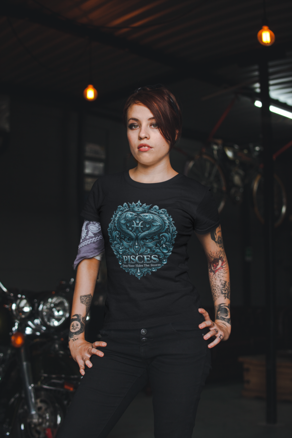 Heavily tattooed female model with an arm band bandana wearing a premium black graphic streetwear t-shirt featuring a detailed dark teal Pisces crest design by Essentia Collective inside a gritty industrial garage.