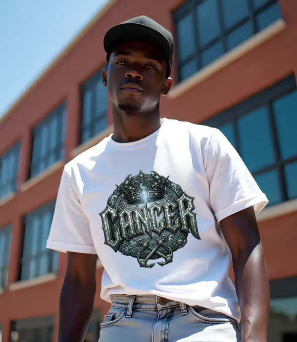 Male model with a black baseball cap looking down wearing a premium white graphic streetwear t-shirt featuring a detailed metallic silver Cancer crown and anchor design by Essentia Collective against a brick industrial building.