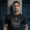 male model leaning against a raw concrete pillar in a dark industrial structure, wearing a black premium Essentia Collective graphic streetwear t-shirt with a high-contrast silver gothic arch design reading Still Standing.