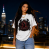 Long-haired female model wearing a gold necklace standing on an urban rooftop at night against a blurred city skyline, sporting a premium white graphic streetwear t-shirt featuring an intricate, gothic Scorpio emblem design by Essentia Collective.
