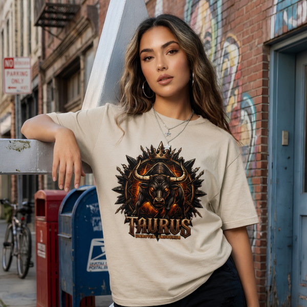 Female model with long wavy light brown hair wearing a silver chain necklace standing on a city street next to a blue mailbox, wearing a premium sand graphic streetwear t-shirt featuring a detailed Taurus bull and crown design by Essentia Collective.