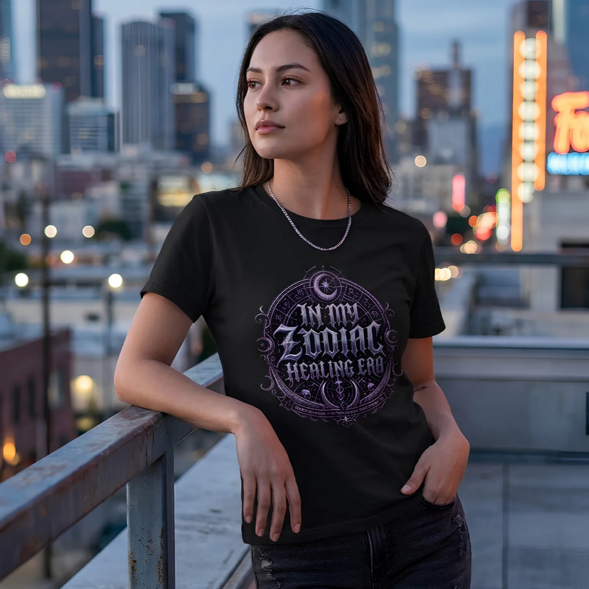 female model leaning on a balcony railing of a metropolitan rooftop during twilight, wearing a black premium graphic t-shirt with an intricate gothic zodiac shield reading In My Zodiac Healing Era.