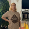 female model with curly blonde hair tied back, standing outside an urban storefront at night, wearing a premium earth-tone mocha tan graphic streetwear t-shirt with a detailed Keep Going emblem design by Essentia Collective.