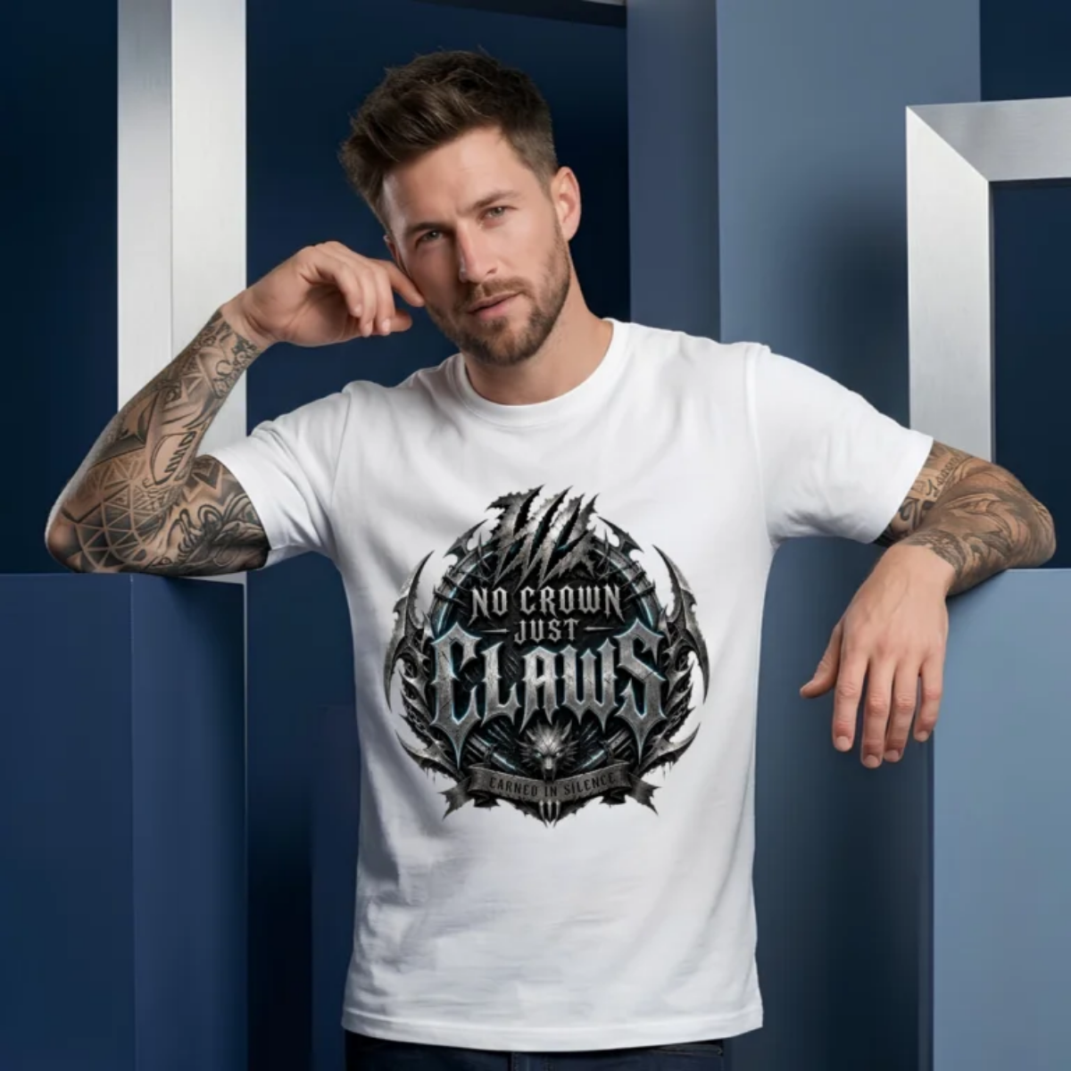 Tattooed male model leaning back against blue and silver geometric column pillars, wearing a white Essentia Collective graphic streetwear t-shirt with a large chiseled silver gothic emblem reading No Crown Just Claws.
