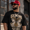 Bearded male model wearing a red cap and black Gemini graphic T-shirt beside an industrial brick building.