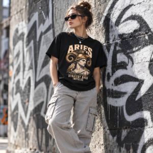 Female model wearing a black Aries graphic T-shirt and gray pants against an urban graffiti-covered wall.